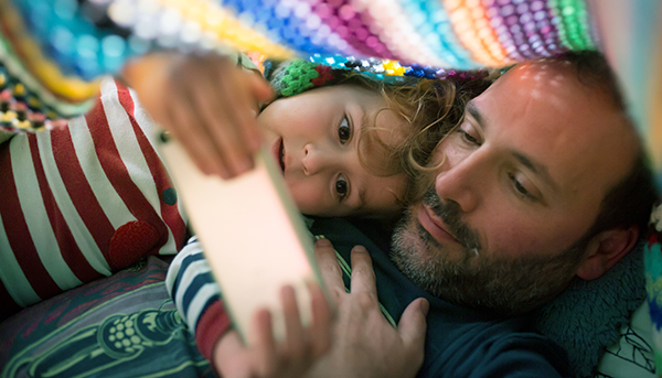 A close up image of a father and child looking at a mobile phone. A colourful blanket is draped over them.