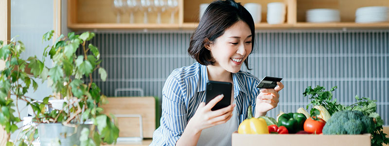 Woman in kitchen holding phone and credit card, representing convenient online banking and secure payment options available through Teachers Mutual Bank, highlighting the ease of managing finances from home.