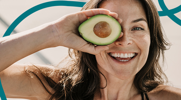 A woman holds half an avocado in front of her eye.