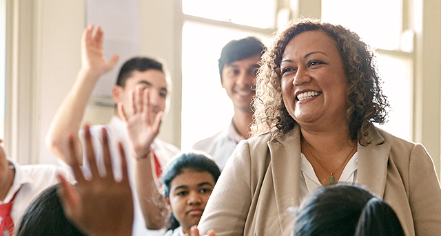 A teacher smiles at her students who are surrounding her, raising their hands.