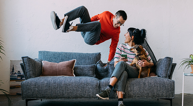 A young man leaps onto his lounge while his significant other sits holding their pet dog.
