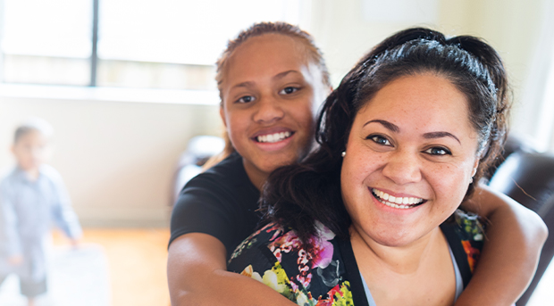 A daughter hugs her mother from behind as both smile at the camera.