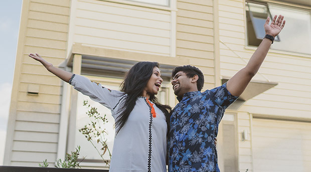 Two first time home owners, a young woman in a pink plaid shirt is piggybacking her partner, a man in a blue business shirt. Both are smiling and the young woman holds the keys to their first home. A kitchen is visible behind them.