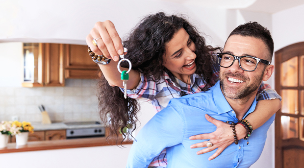 Two first time home owners, a young woman in a pink plaid shirt is piggybacking her partner, a man in a blue business shirt. Both are smiling and the young woman holds the keys to their first home. A kitchen is visible behind them.