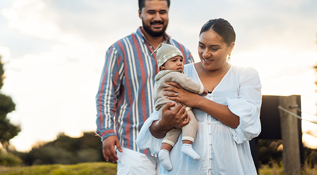 A mother walks through farmland and holds her child while the child's father follows behind.