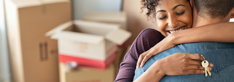 A home buyer hugs her partner and smiles with joy, holding the keys to their property in her hand.