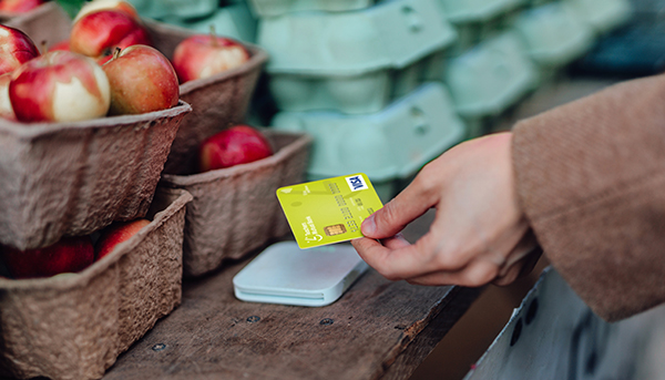 A close up of a hand tapping a Teachers Mutual Bank lime green credit card onto a card reader. In the background are cartons of eggs and apples.