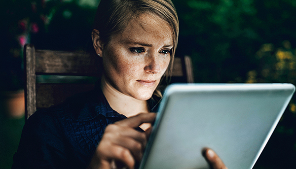 A close up image of a serious-looking woman using a tablet device while sitting in a chair. The background is dark.