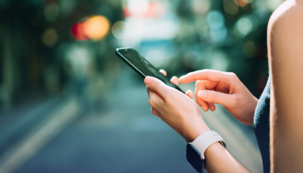 A close up image of a hand wearing a digital watch and holding a mobile phone. Blurred trees and the street are visible in the background.