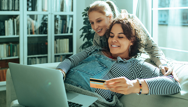 A woman holding a bank card lies on a couch with her partner leaning over her shoulder. Both are looking at a laptop in front of them. Bookshelves and a window are behind them.