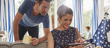 A man and woman smiling and looking at a tablet while children play in the background