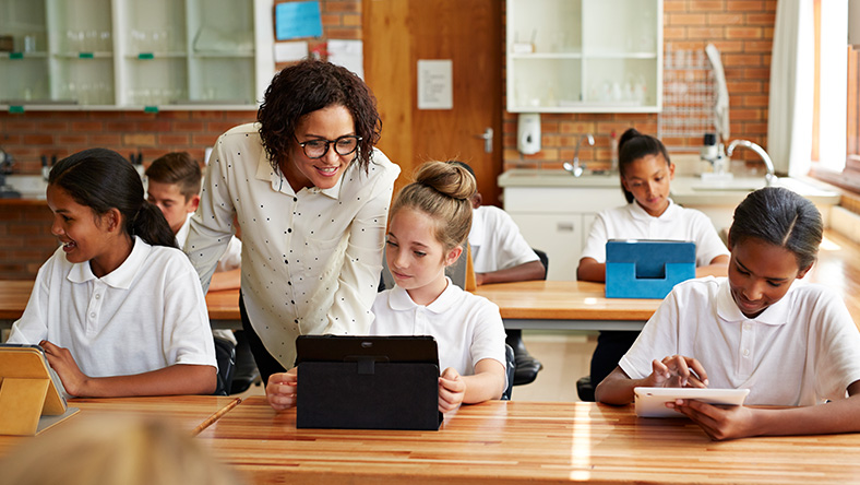 Female teacher in a room filled with students, looking at a young female students' laptop screen