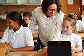 Female teacher in a room filled with students, looking at a young female students' laptop screen