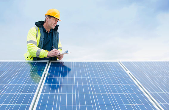 A construction worker wearing a hard hat and yellow vest, writing notes while assessing solar panels