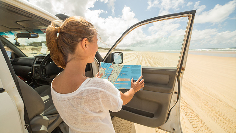 A woman stands among sand dunes next to her car with the door open, reading a map.