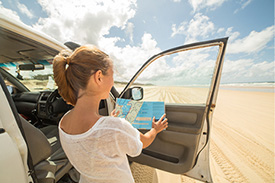 A woman stands among sand dunes next to her car with the door open, reading a map.