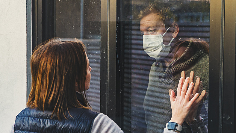 A man in a medical mask holds his hand to a woman through a window, practicing caremongering.