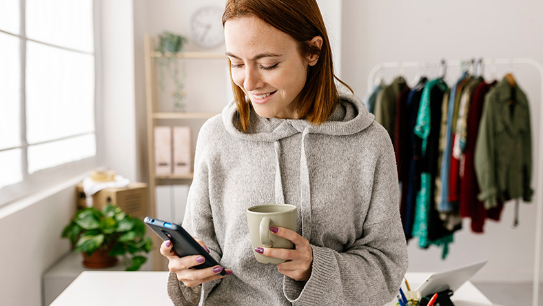 A woman looks at her phone in her right hand holding a mug in her other hand.