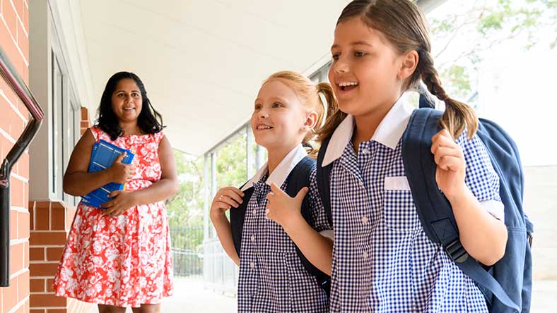 Two children wearing backpacks, looking excited in school environment with teacher in a pink dress watches on