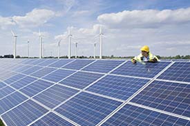 Worker wearing high visibility work wear climbing on a roof covered in solar panels in front of a wind farm