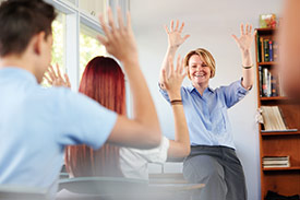 Teacher at front of a class room encouraging students to raise their hands. 