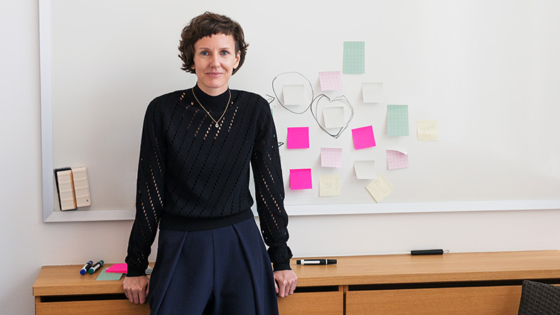 A young woman in business attire leaning on desk with a whiteboard behind her and post-it notes stuck to the board