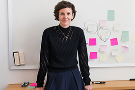 A young woman in business attire leaning on desk with a whiteboard behind her and post-it notes stuck to the board