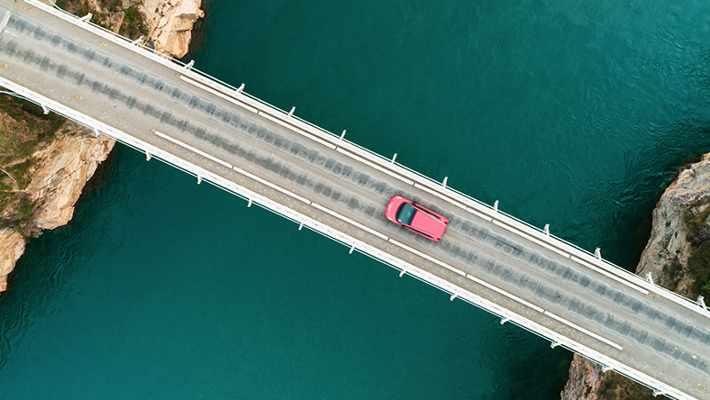 A birds eye view shot shows a new red car driving across a bridge over a large lake.