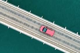 A birds eye view shot shows a new red car driving across a bridge over a large lake.