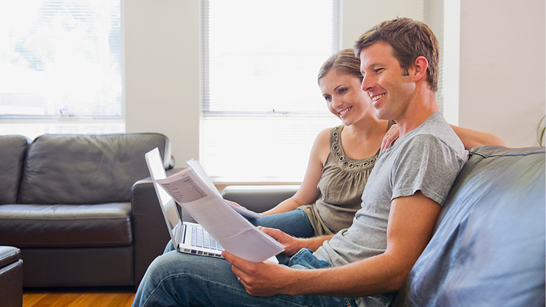 A couple sit on the couch in their home, reading documents to check their financial wellness.