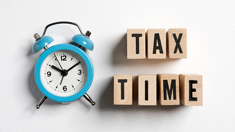 Image of a blue alarm clock laid flat next to wooden building blocks with letters spelling out 'Tax Time'.