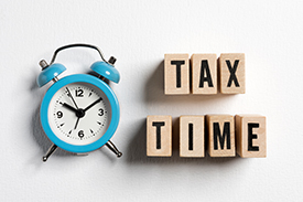 Image of a blue alarm clock laid flat next to wooden building blocks with letters spelling out 'Tax Time'.