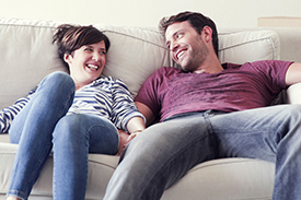 A happy pair of first home buyers lounge on the couch and smile at each other. 5 must read tips for first home buyers.