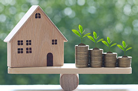 A wooden toy house balances on a set of scales next to four piles of coins.