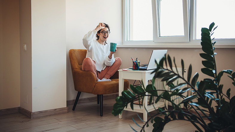 A happy young woman researches home loans on an armchair while sipping coffee