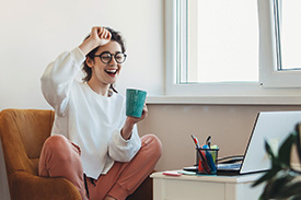 A happy young woman researches home loans on an armchair while sipping coffee