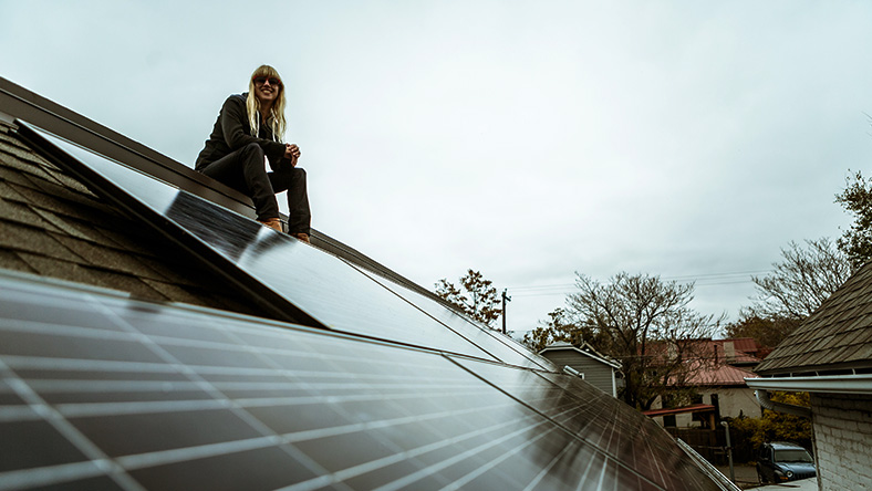 A young woman sits on a rooftop next to her solar panel grid observing the view.