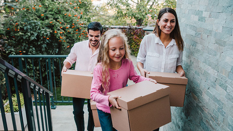 A young smiling family carry boxes into their new home.