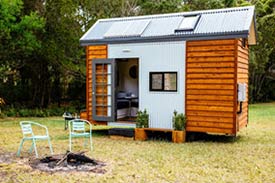 Image of a tree surrounded tiny house with wood paneling and skylights and a fire pit with a couple of chairs in the front