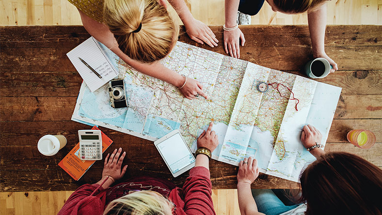 Four friends standing around a map of the world plotting their next holiday destination