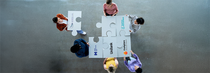 Five people holding the logos of the TMBL brands as puzzle pieces with the logo of Australian Mutual Bank as another puzzle piece joining.