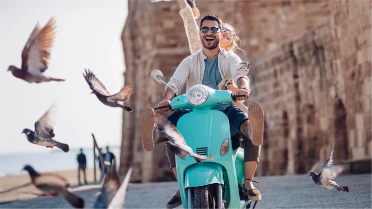 A young couple rides a bright turquoise scooter along a cobblestone street, surrounded by flying pigeons.