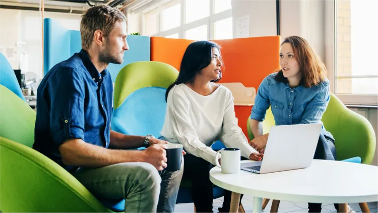 Three adults are engaged in a discussion around a white table in a contemporary office lounge.