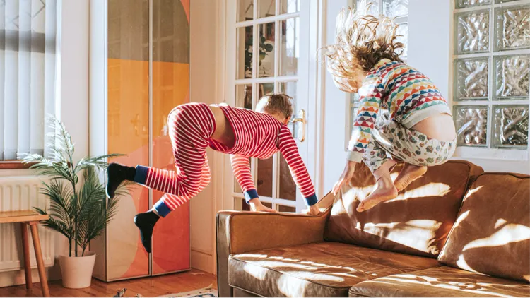 Two children in colorful pajamas are energetically jumping on a brown couch in a bright, sunlit living room.