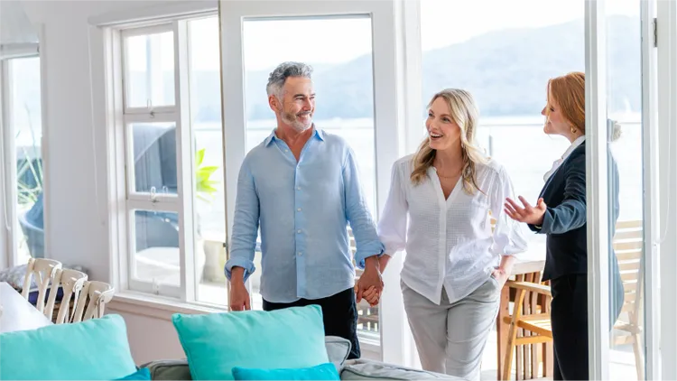A middle-aged couple is shown holding hands while touring a bright, modern home with large windows and waterfront.