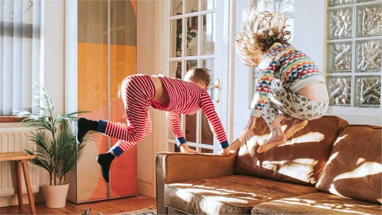 Two children in colorful pajamas are energetically jumping on a brown couch in a bright, sunlit living room.