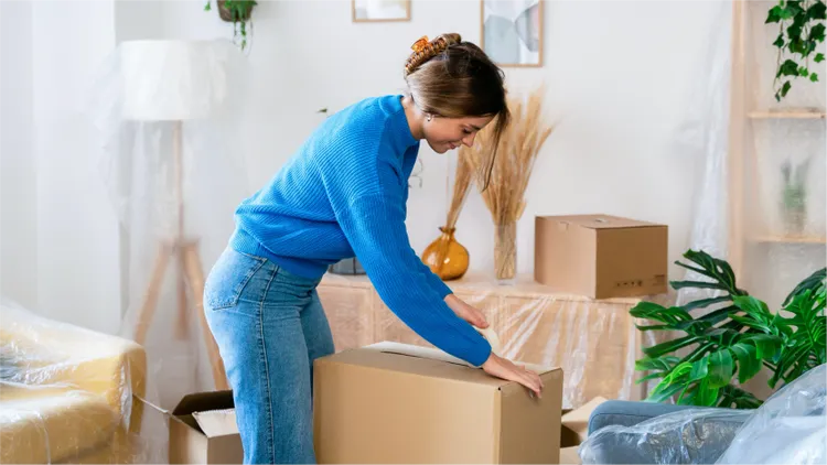 A young woman in a blue sweater is seen packing a cardboard box in a living room.