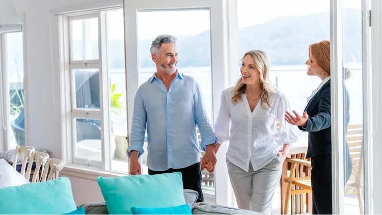 A middle-aged couple is shown holding hands while touring a bright, modern home with large windows and waterfront.