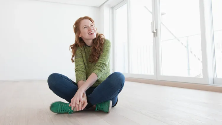 A young adult woman with long red hair sits cross-legged on a light wooden floor.