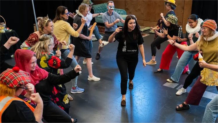 A diverse group of adults participates in a lively theater rehearsal, forming a circle with dramatic gestures.